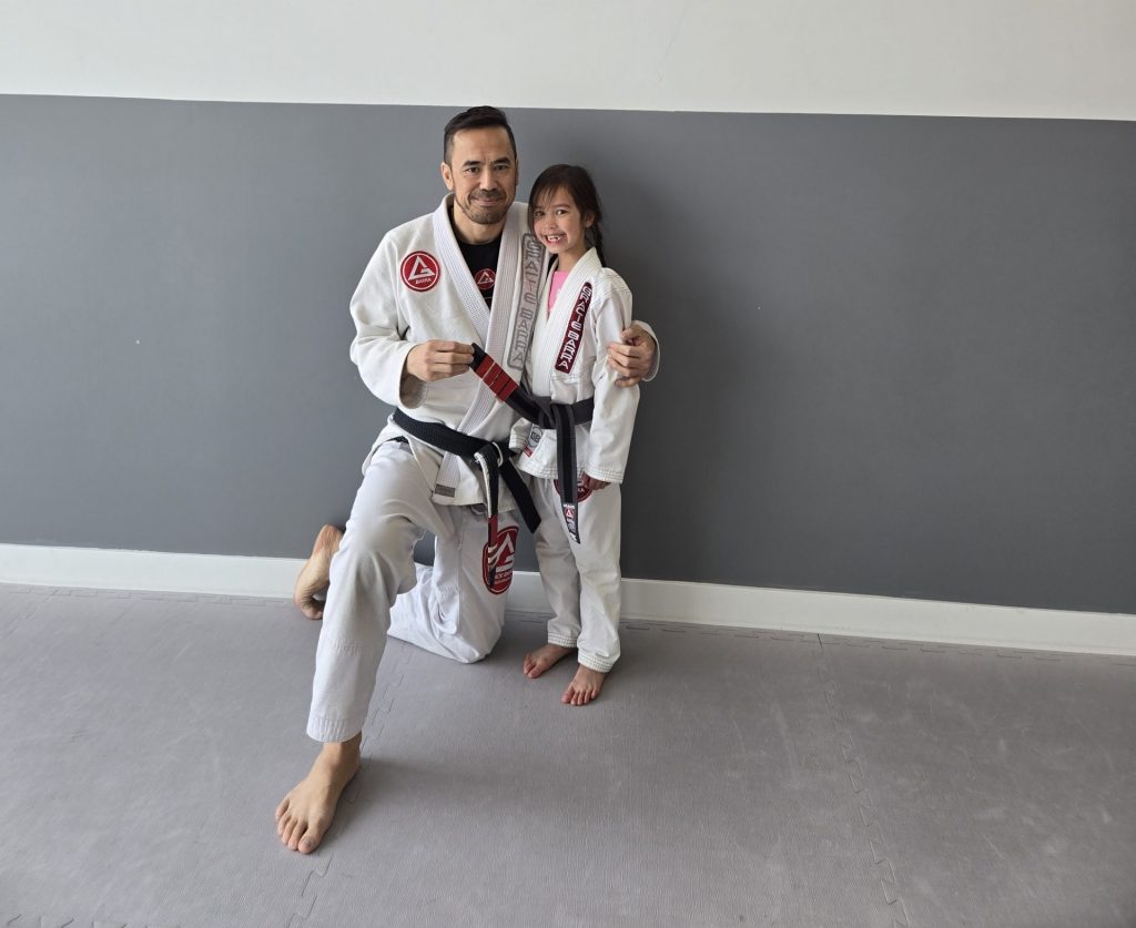 A martial arts instructor kneels beside his young daughter, both wearing Gracie Barra Jiu-Jitsu uniforms in front of the Gracie Barra Lake Country logo on the wall.