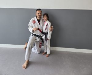 A martial arts instructor kneels beside his young daughter, both wearing Gracie Barra Jiu-Jitsu uniforms in front of the Gracie Barra Lake Country logo on the wall.