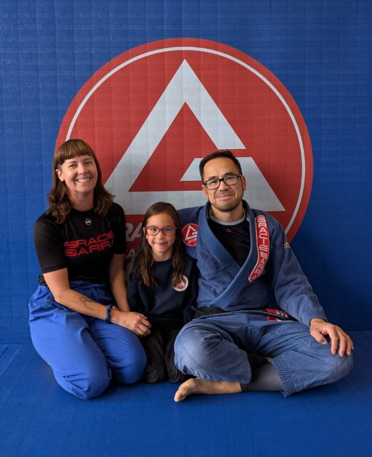 A smiling family of three, sitting on a Jiu-Jitsu mat in front of the Gracie Barra logo. The father wears a blue Gi, the daughter wears a Gracie Barra sweatshirt, and the mother wears a Gracie Barra t-shirt.